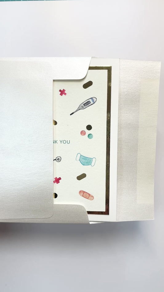 Hands opening an envelope to reveal a handmade healthcare thank you card with medical icons and gold foil.