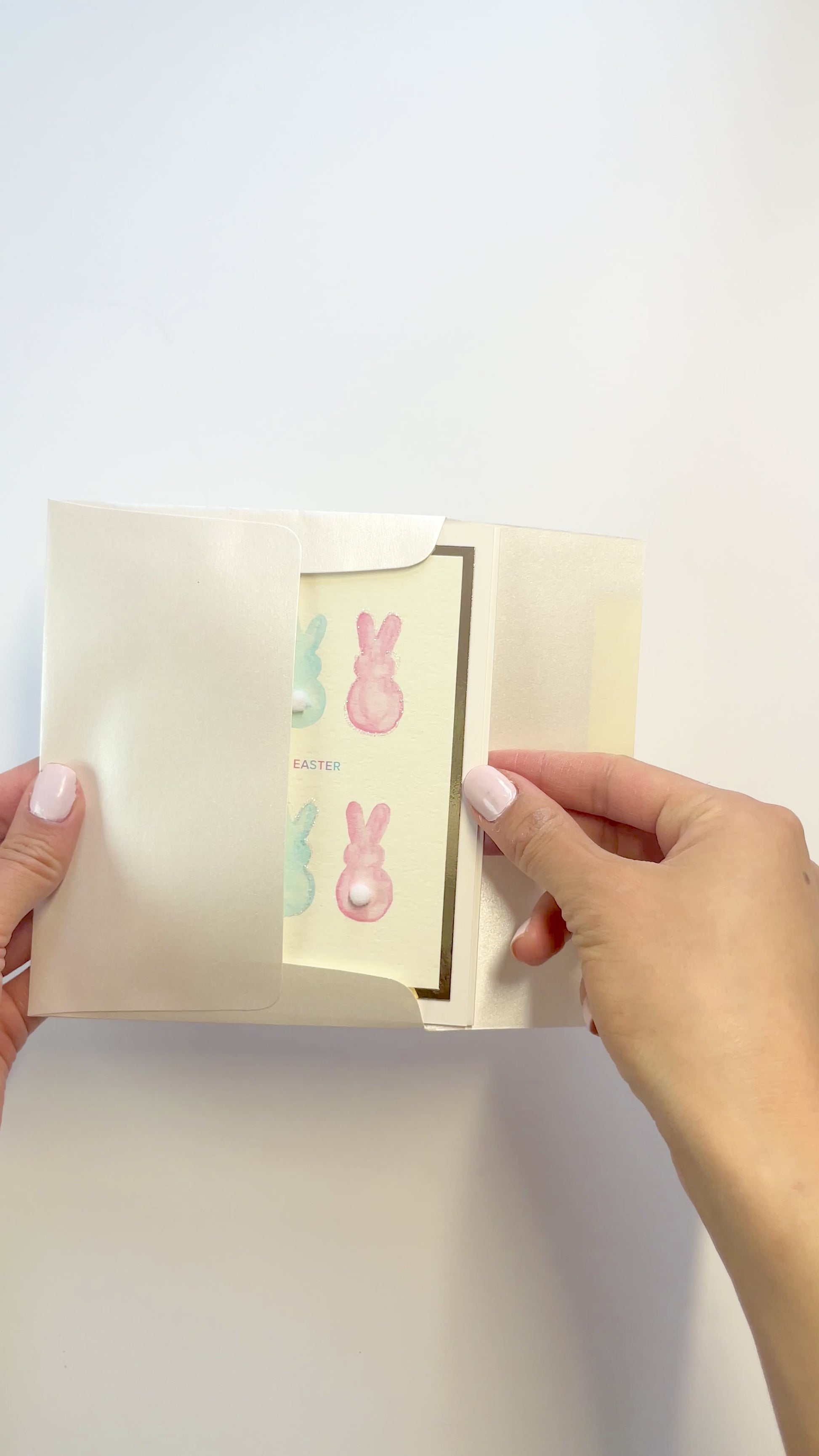 Hands opening an envelope to reveal a handmade Easter card with watercolor bunnies and pom-poms.