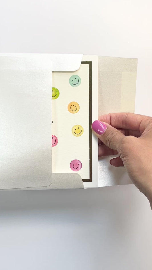 Hands opening an envelope to reveal a handmade hello card with colorful smiley faces and gold foil details.