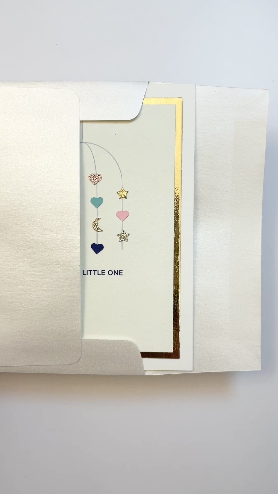 Hands opening an envelope to reveal a handmade multicolor baby card with watercolor mobile artwork and gold foil trim.