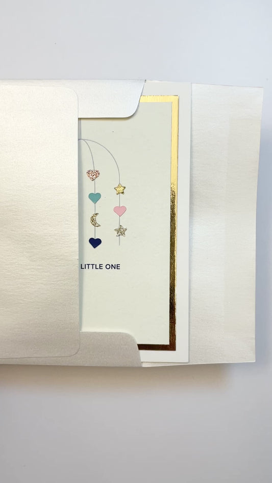 Hands opening an envelope to reveal a handmade multicolor baby card with watercolor mobile artwork and gold foil trim.