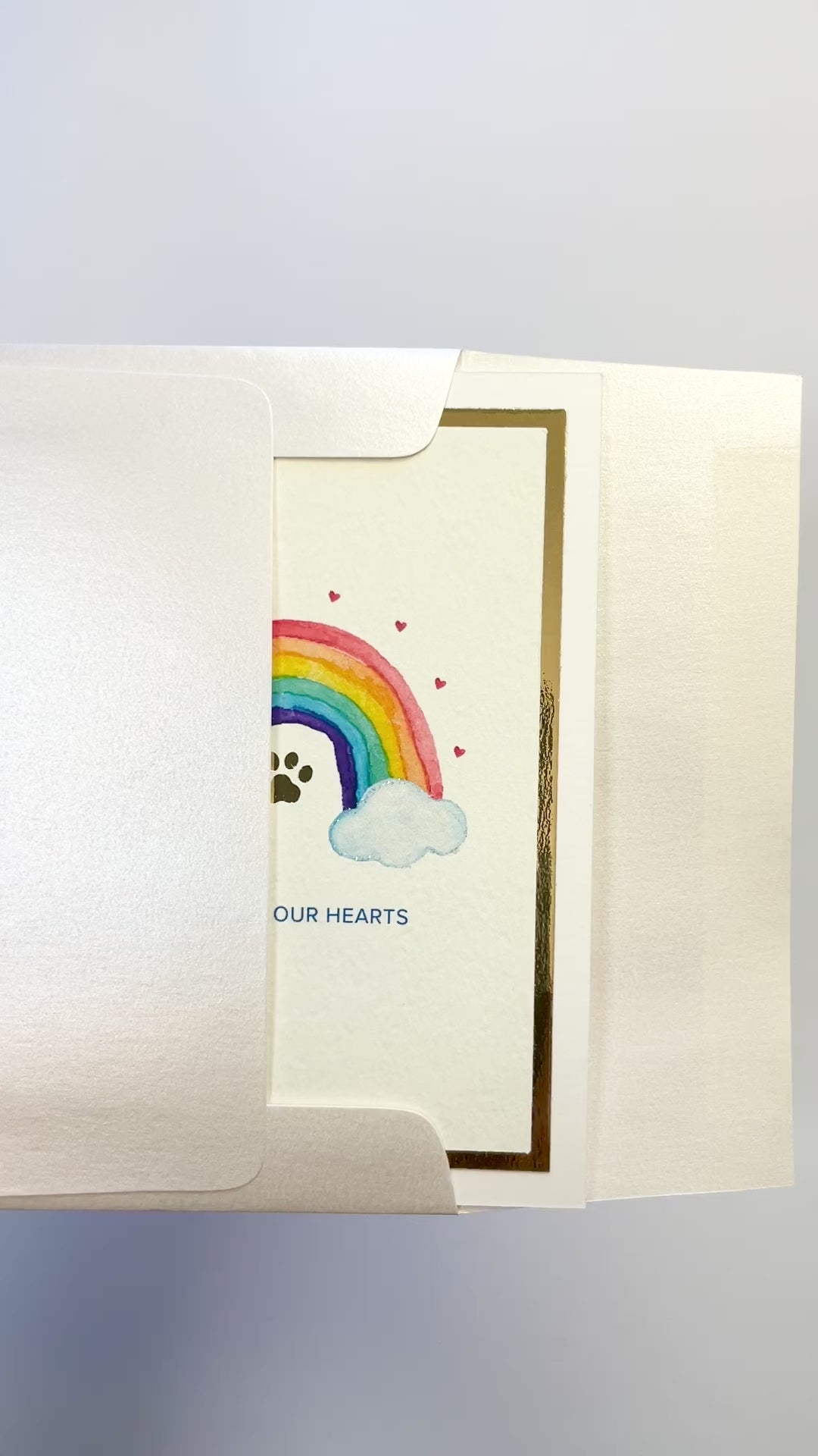 Hands opening an envelope to reveal a handmade pet loss sympathy card with rainbow and gold foil.