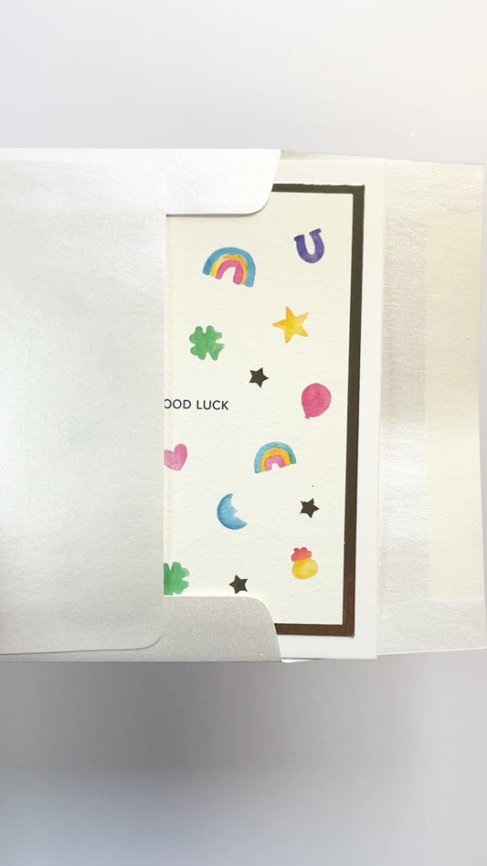 Hands opening an envelope to reveal a handmade good luck charms card with watercolor icons and gold foil.