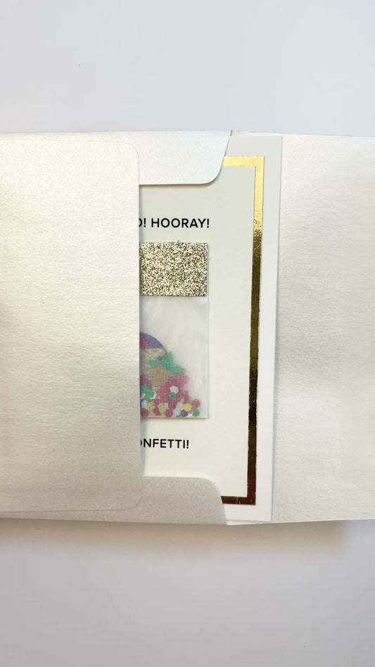 Hands opening an envelope to reveal a handmade confetti celebration card with gold foil and colorful art.