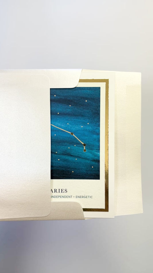 Hands opening an envelope to reveal a handmade Aries zodiac constellation card with gold foil accents.