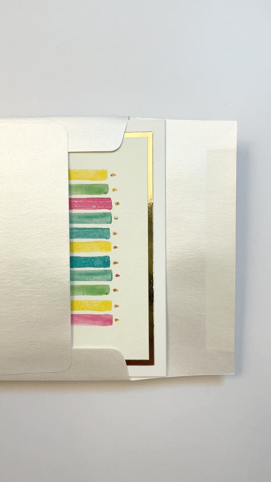 Hands opening an envelope to reveal a handmade multicolor birthday candles card with watercolor art and gold foil accents.