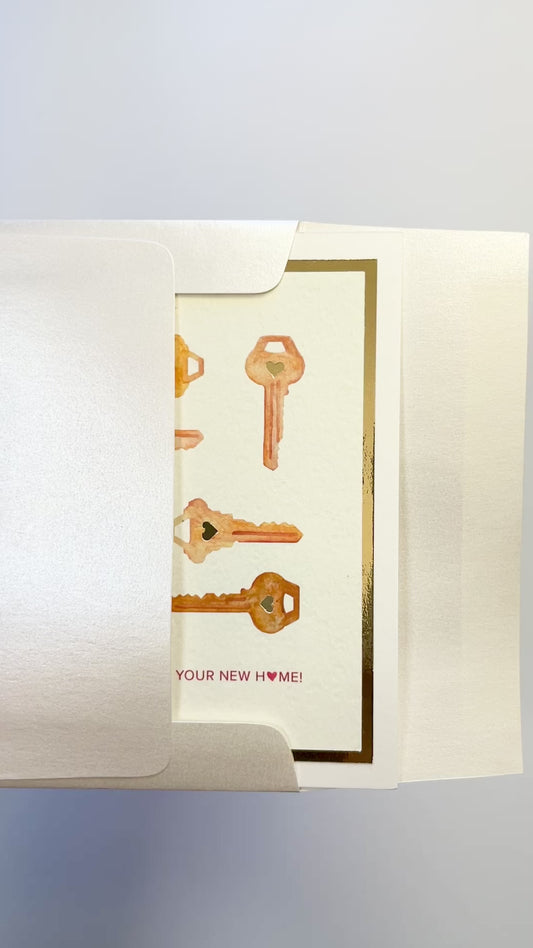 Hands opening an envelope to reveal a handmade housewarming card with gold foil key designs.