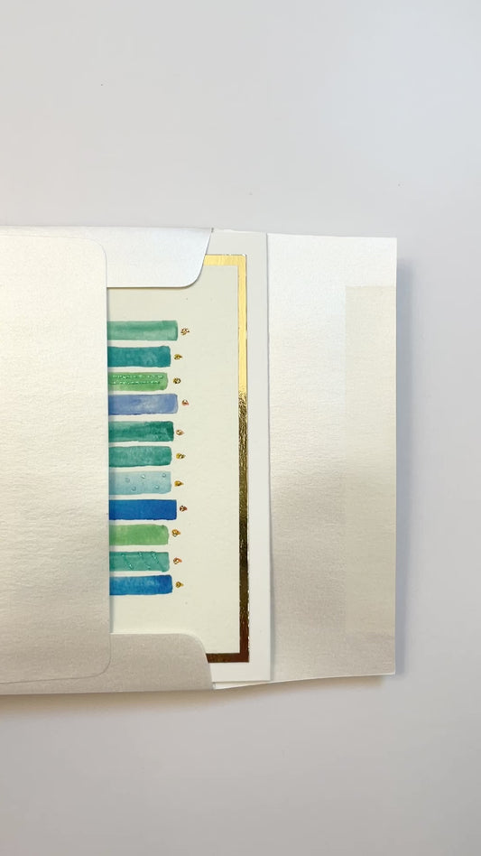 Hands opening an envelope to reveal a handmade blue and green birthday candles card with gold foil details.