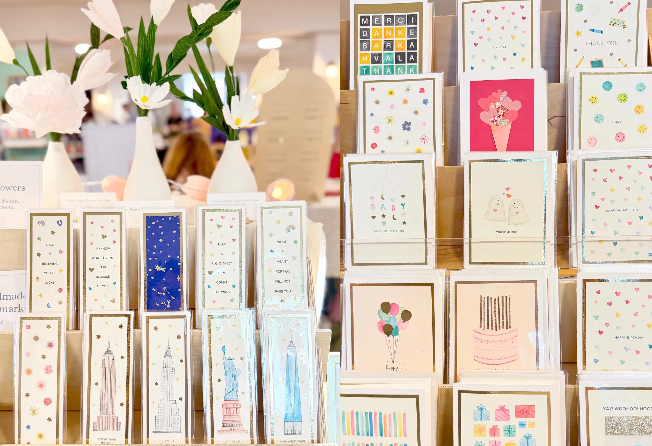 A display of colorful greeting cards arranged on a stand in a shop or event setting, with white flowers in vases in the blurred background.