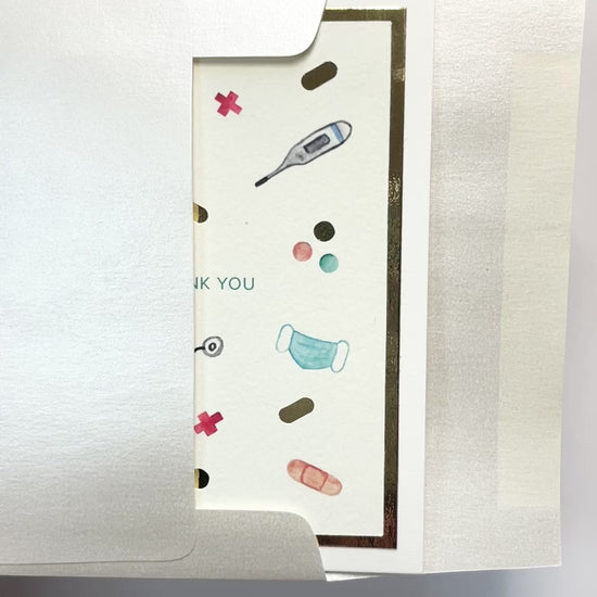 Hands opening an envelope to reveal a handmade healthcare thank you card with medical icons and gold foil.