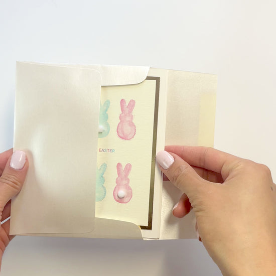 Hands opening an envelope to reveal a handmade Easter card with watercolor bunnies and pom-poms.