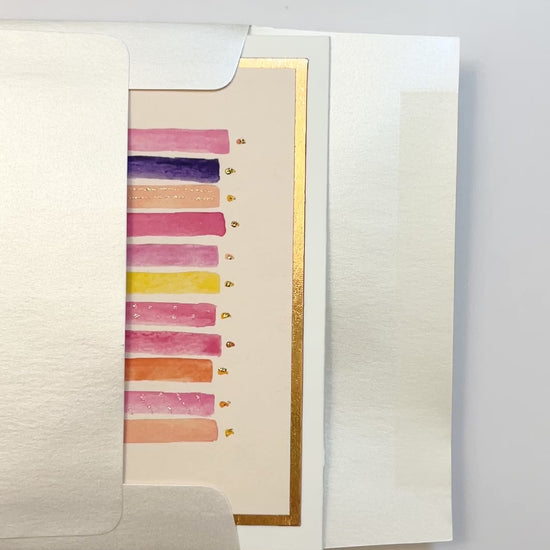 Hands opening an envelope to reveal a handmade pink and purple birthday candles card with rose gold foil.