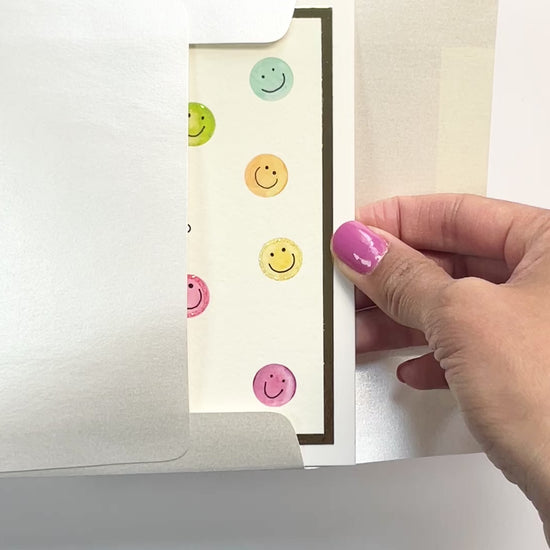 Hands opening an envelope to reveal a handmade hello card with colorful smiley faces and gold foil details.