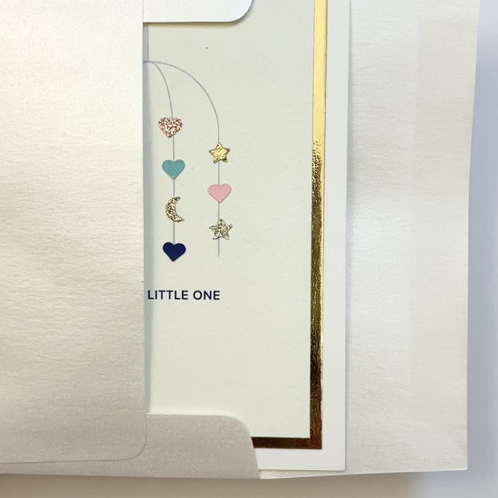 Hands opening an envelope to reveal a handmade multicolor baby card with watercolor mobile artwork and gold foil trim.
