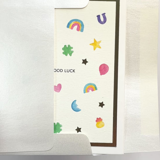 Hands opening an envelope to reveal a handmade good luck charms card with watercolor icons and gold foil.