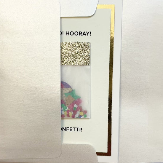 Hands opening an envelope to reveal a handmade confetti celebration card with gold foil and colorful art.