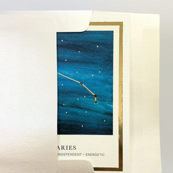 Hands opening an envelope to reveal a handmade Aries zodiac constellation card with gold foil accents.