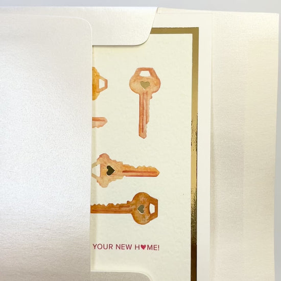 Hands opening an envelope to reveal a handmade housewarming card with gold foil key designs.