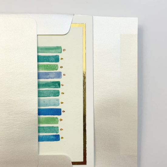 Hands opening an envelope to reveal a handmade blue and green birthday candles card with gold foil details.